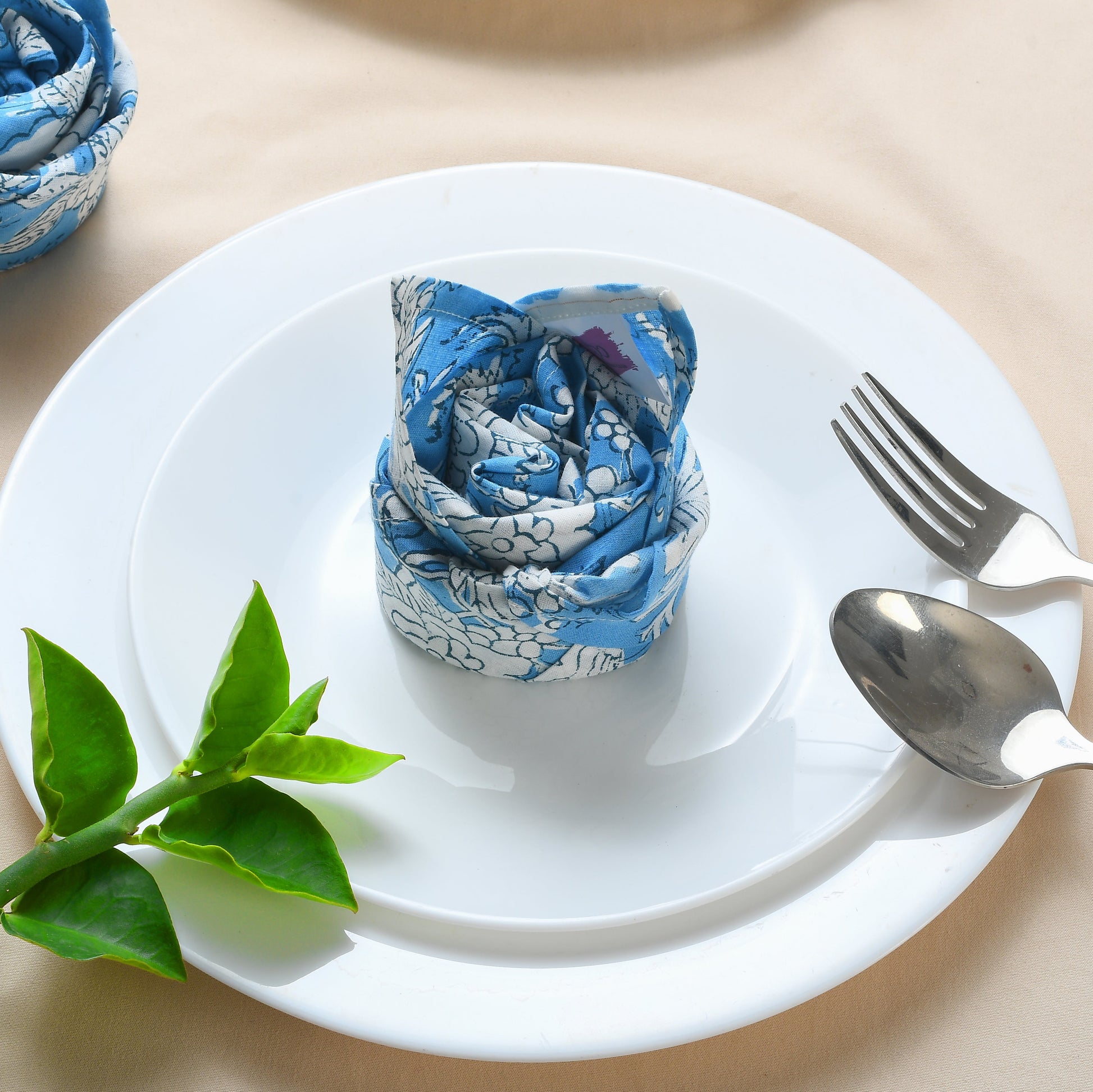 Rolled blue floral napkin placed upright on a plate, styled with green leaves and silverware.
