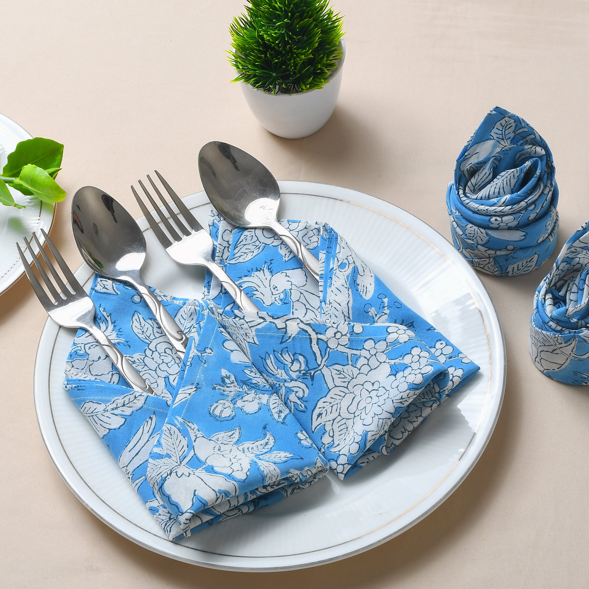Place setting with blue floral napkin tucked under silverware on a white plate and napkins rolled nearby.