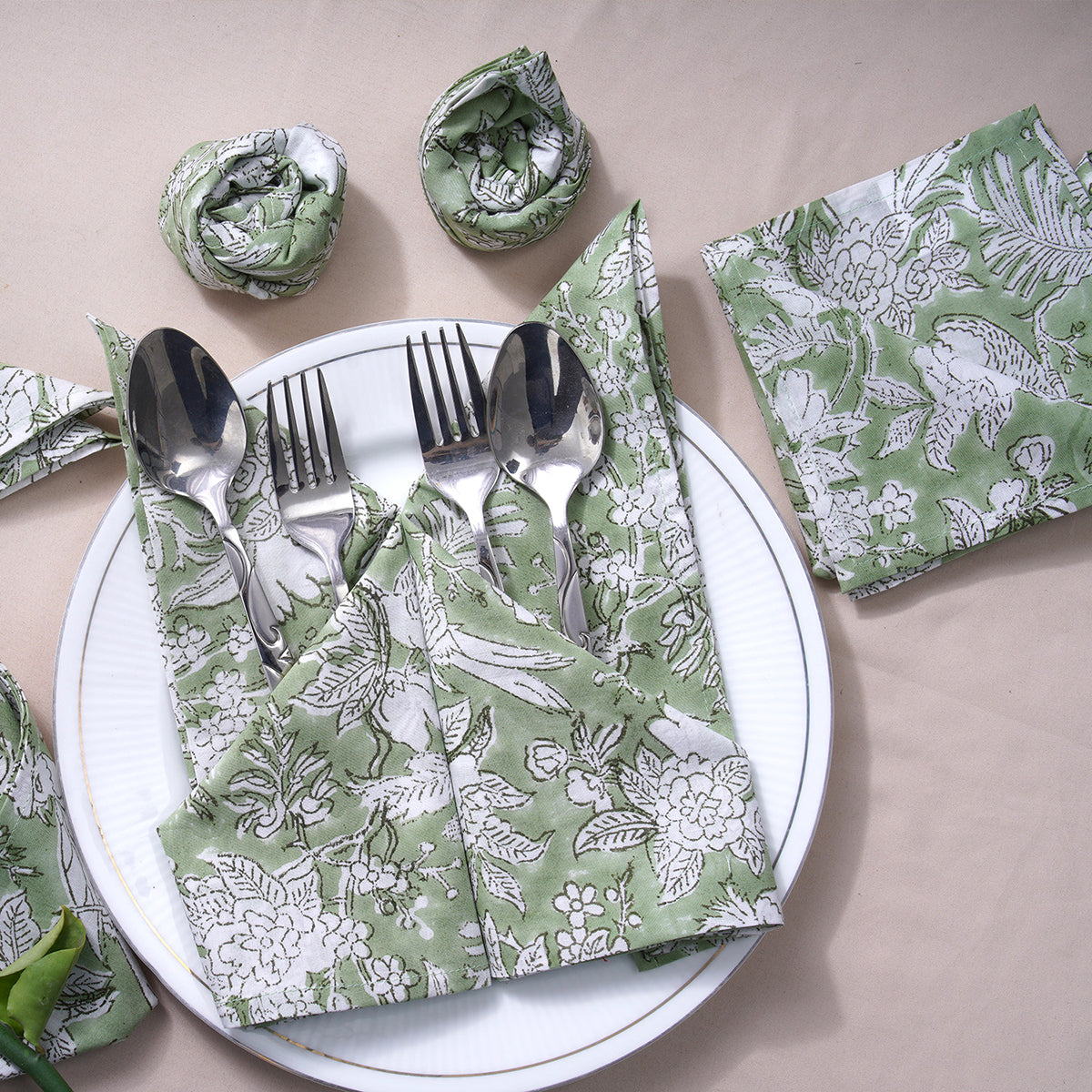 A set of olive green cotton cloth napkins with floral and geometric patterns, displayed on a white plate with cutlery and a matching napkin ring.