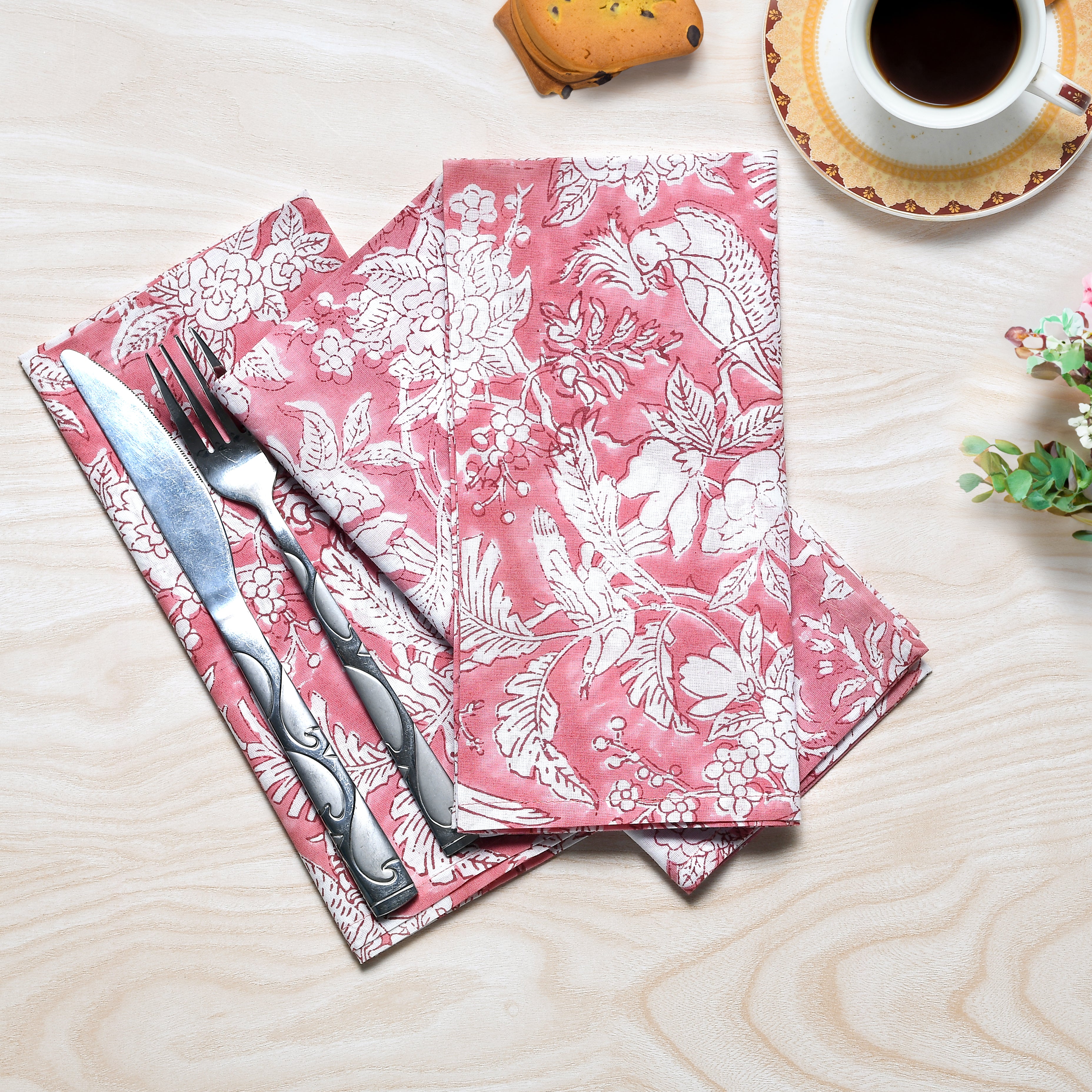 A set of coral pink cotton cloth napkins with floral and geometric hand-printed designs, displayed on a wooden table alongside a fork, knife, and a cup of coffee.