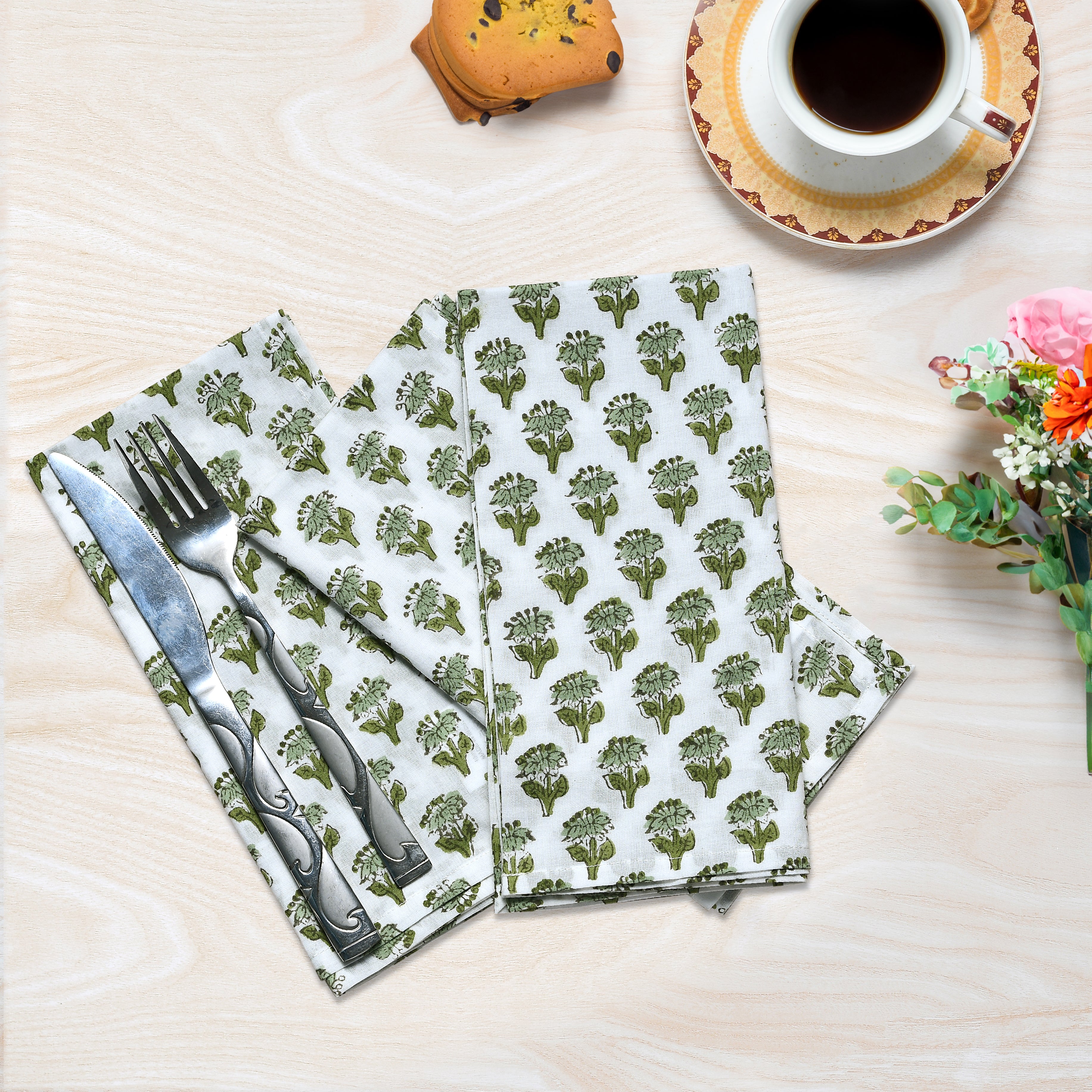 A set of emerald green floral patterned cotton cloth napkins displayed on a wooden table alongside a cup of coffee and decorative flowers.