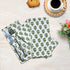A set of emerald green floral patterned cotton cloth napkins displayed on a wooden table alongside a cup of coffee and decorative flowers.