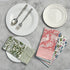 A collection of colorful cotton cloth napkins with striped border, displayed on a table alongside white plates and cutlery.
