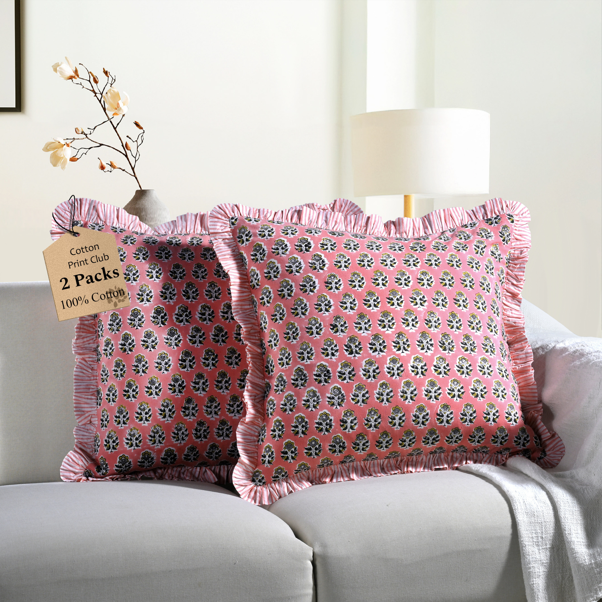 Two pink floral block print throw pillows with ruffled edges on a white sofa beside a modern lamp.