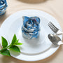 Rolled blue floral napkin placed upright on a plate, styled with green leaves and silverware.

