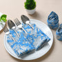 Place setting with blue floral napkin tucked under silverware on a white plate and napkins rolled nearby.

