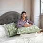 Woman sitting on a bed with green floral pillows holding a cup.