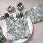 A set of olive green cotton cloth napkins with floral and geometric patterns, displayed on a white plate with cutlery and a matching napkin ring.