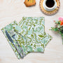 A set of green cotton cloth napkins with floral and geometric hand-printed patterns, displayed on a wooden surface with a fork, knife, and a cup of coffee in the background.