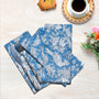 A set of blue cotton cloth napkins with a white floral and geometric pattern, displayed on a table with a cup of coffee and decorative flowers in the background.
