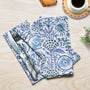 A set of folded indigo blue cotton cloth napkins with floral and geometric patterns, displayed on a wooden table with a fork, knife, and a cup of coffee in the background.