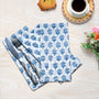 A set of blue and white floral patterned cotton cloth napkins displayed on a wooden surface with a fork, knife, and a cup of coffee in the background.