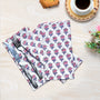 A pair of folded cotton cloth napkins with a floral pattern, displayed on a wooden surface alongside a fork, knife, and a cup of coffee.