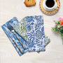 A set of folded cotton cloth napkins with floral and geometric patterns, displayed on a table accompanied by cutlery and a cup of coffee.
