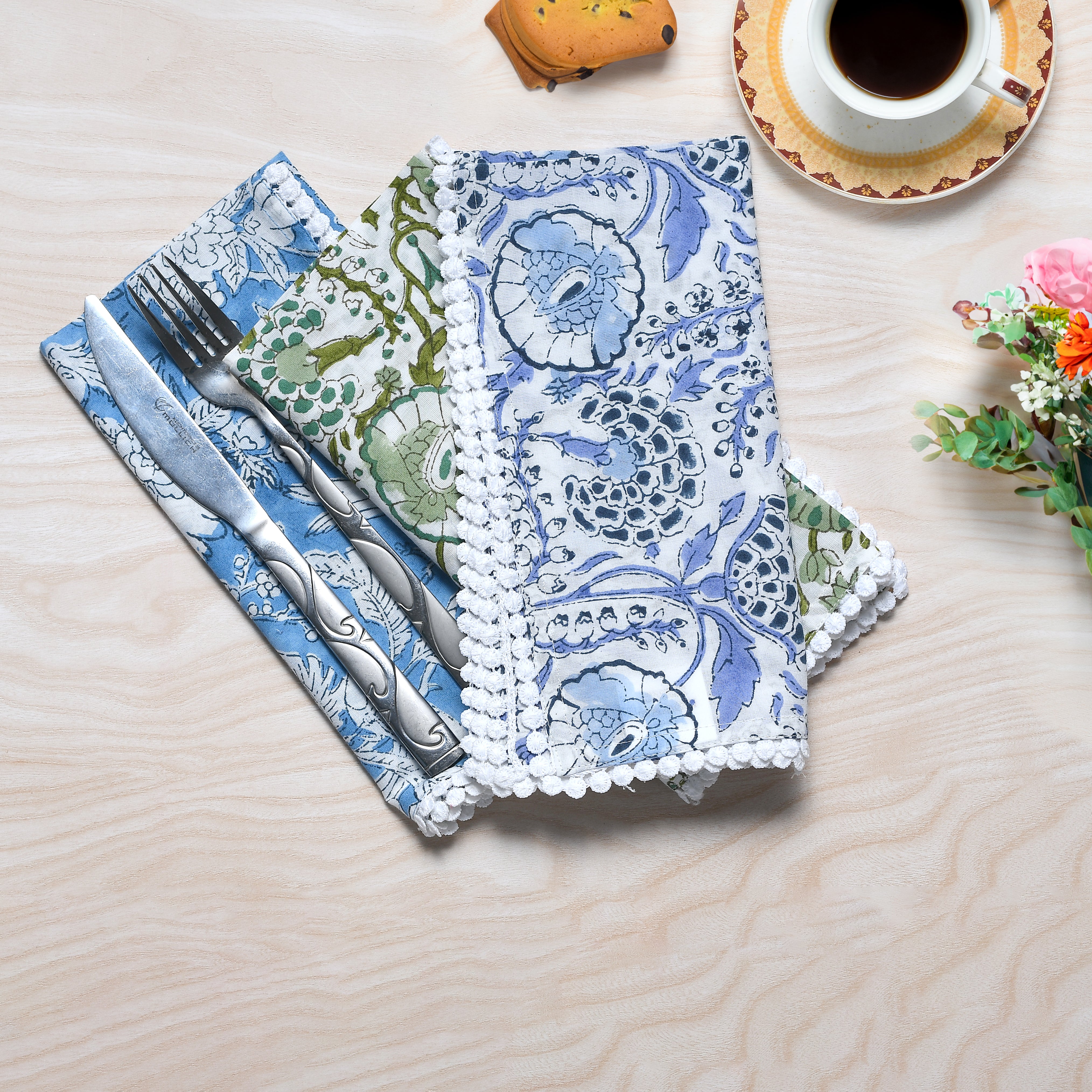 A set of folded cotton cloth napkins with a floral pattern, displayed on a wooden surface, accompanied by a fork, knife, and a cup of coffee in the background.