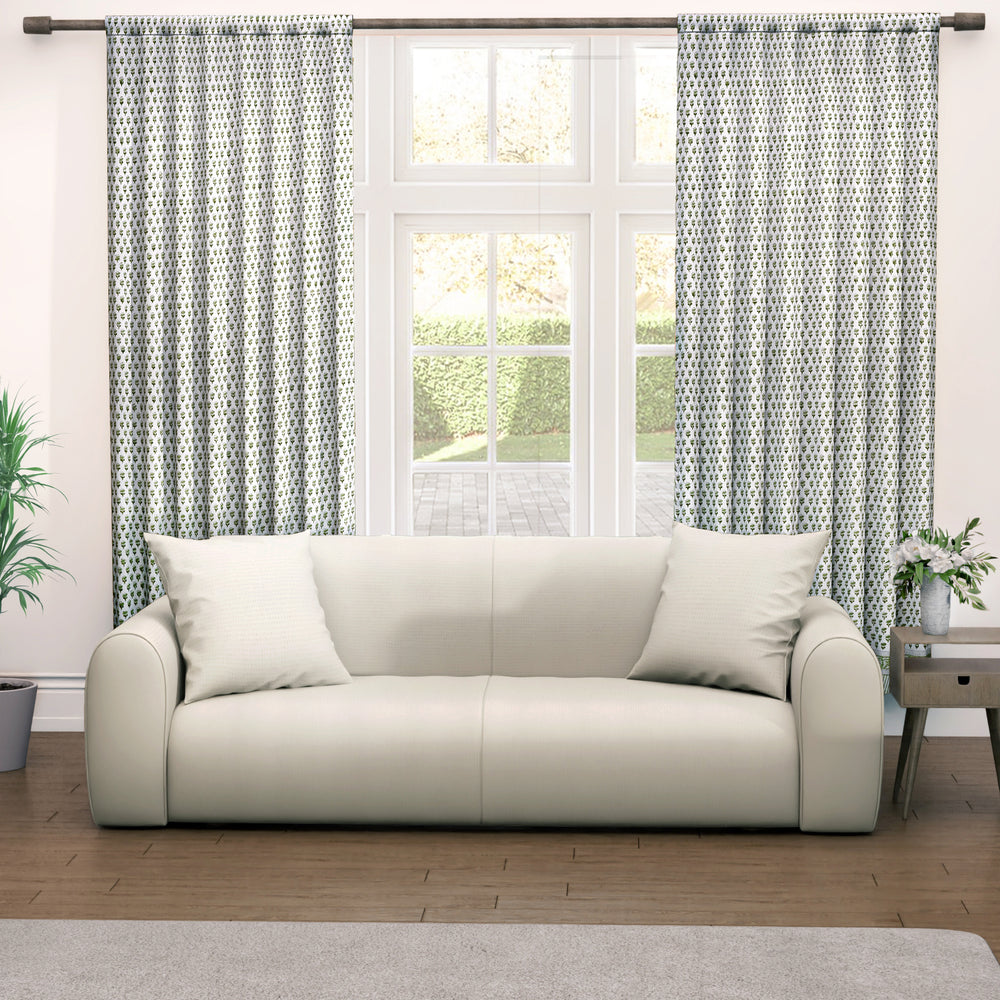 Semi-sheer cotton block print curtains in emerald green, hanging on a rod pocket, allowing natural light to filter through, with a patterned design, placed in a room with a chair and a plant.