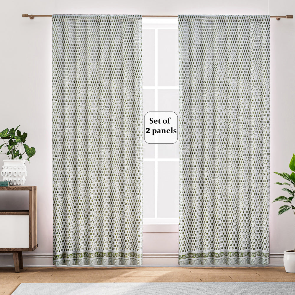 Semi-sheer cotton block print curtains in emerald green, hanging on a rod pocket, allowing natural light to filter through, with a patterned design, placed in a room with a chair and a plant.