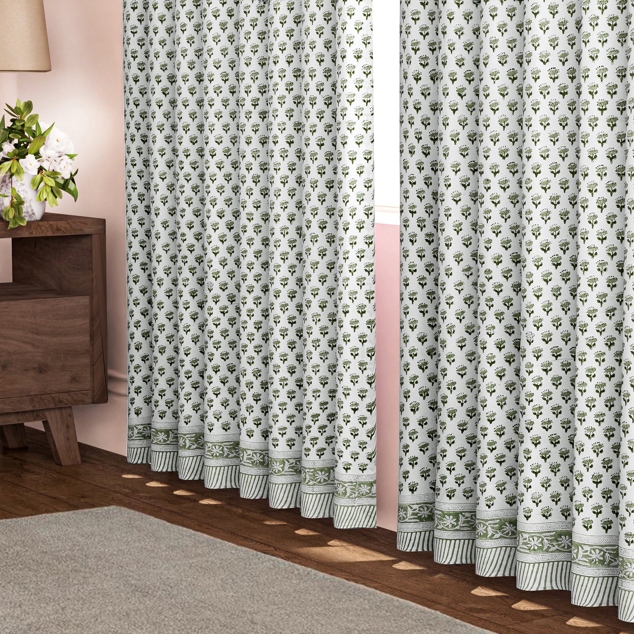 Semi-sheer cotton block print curtains in emerald green, hanging on a rod pocket, allowing natural light to filter through, with a patterned design, placed in a room with a chair and a plant.