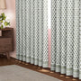 Semi-sheer cotton block print curtains in emerald green, hanging on a rod pocket, allowing natural light to filter through, with a patterned design, placed in a room with a chair and a plant.