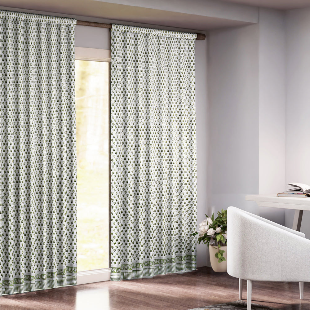 Semi-sheer cotton block print curtains in emerald green, hanging on a rod pocket, allowing natural light to filter through, with a patterned design, placed in a room with a chair and a plant.