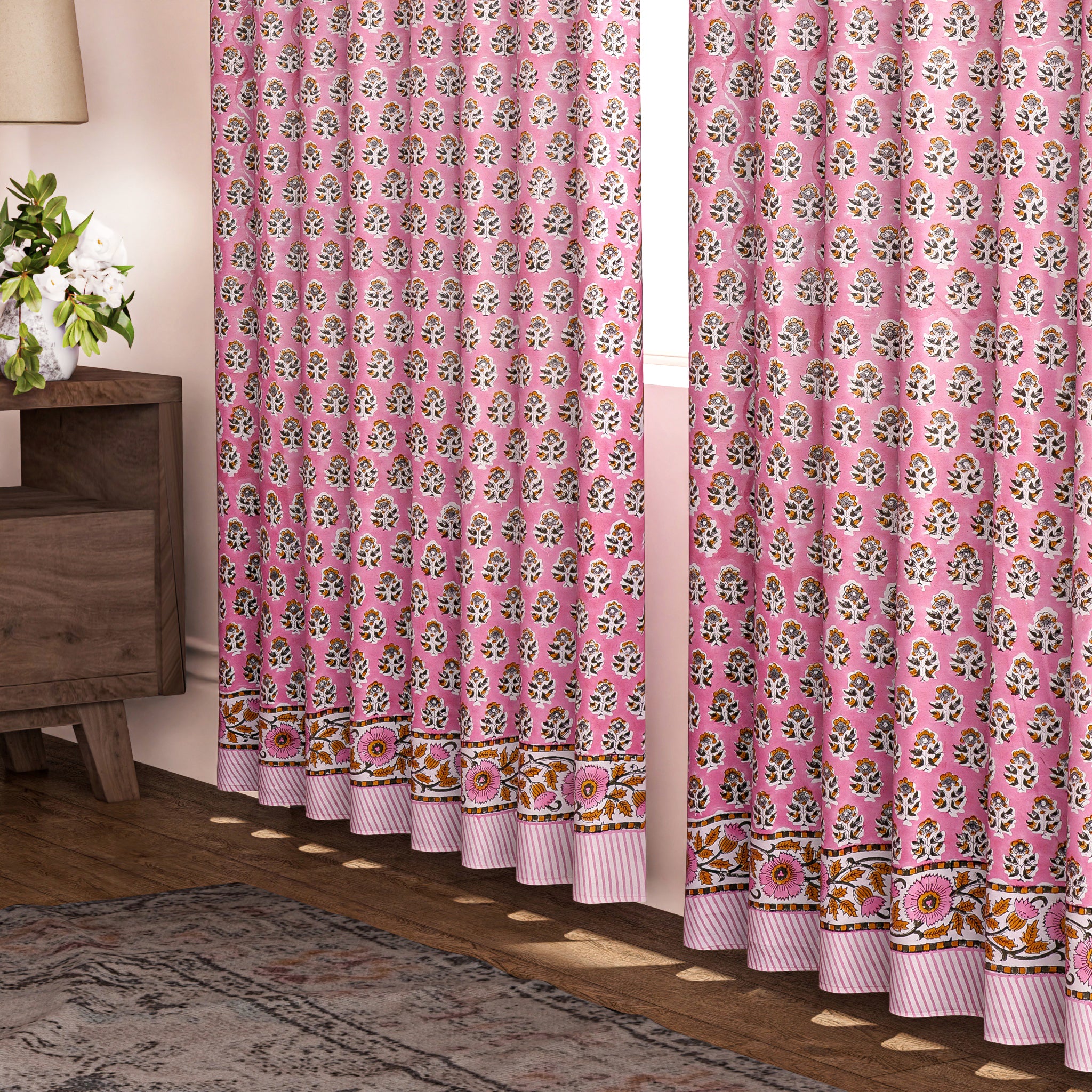 Semi-sheer cotton block print curtains in poppy pink with intricate patterns, hanging in a room with a light filtering through.